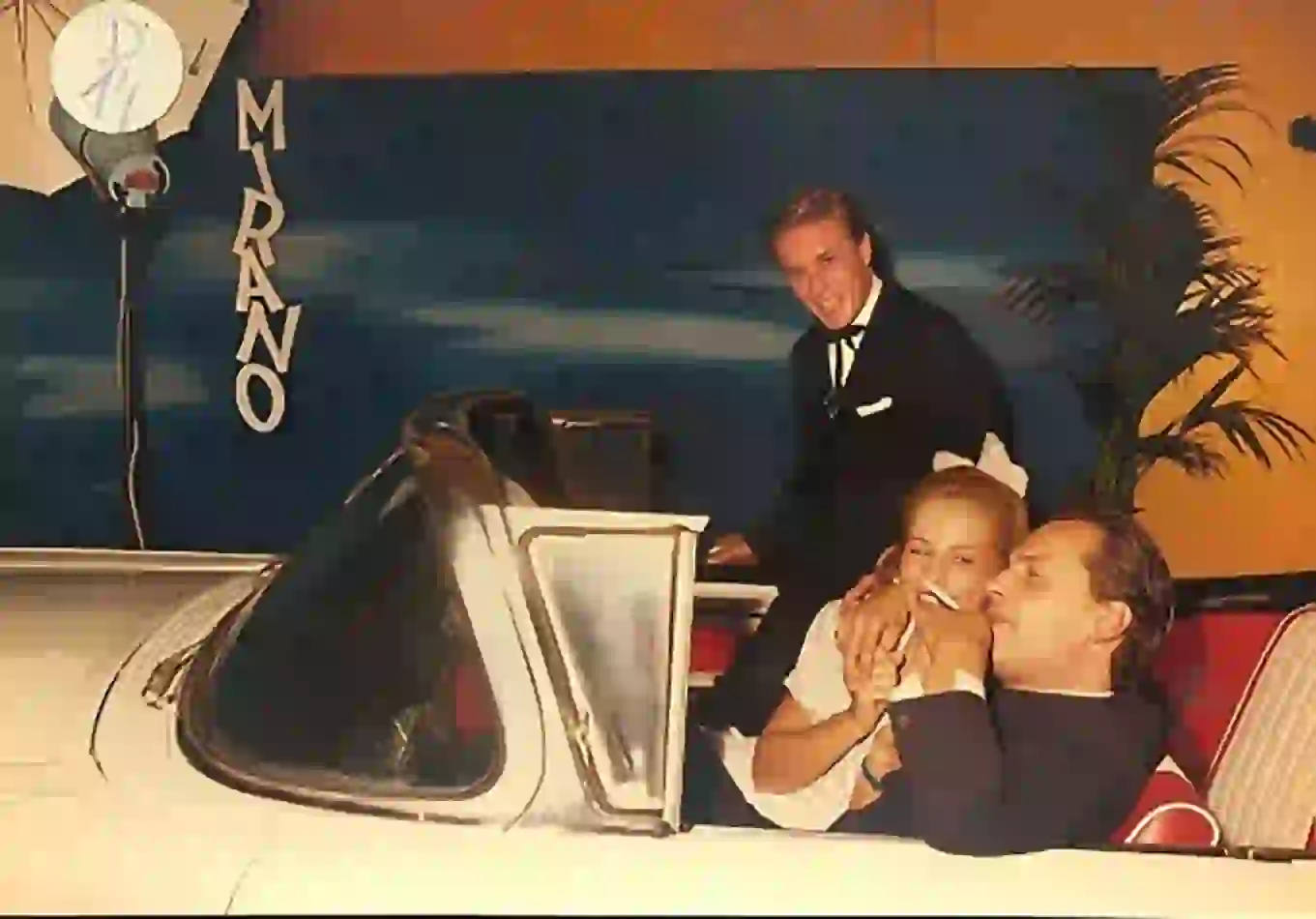 Photographie couleur, format horizontal. Dans le hall d'entrée du Mirano Continental, une Cadillac blanche est aménagée comme point de rendez-vous : un homme en smoking et deux femmes élégantes posent à bord, devant un décor bleu nuit et le logo vertical du club. Années 1980.