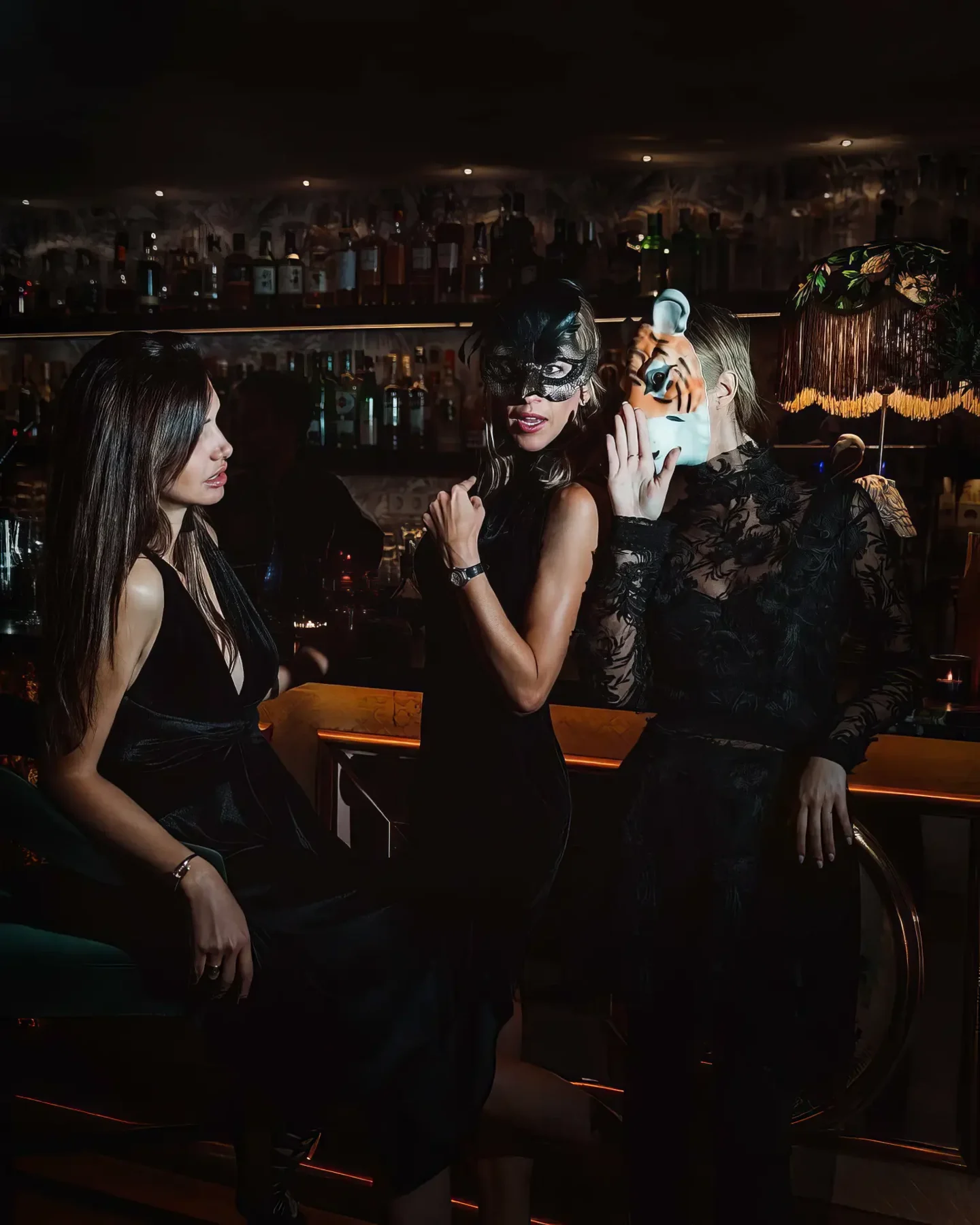 Trois silhouettes en noir devant le mur de bouteilles, masque de plumes et masque de tigre à la main. Le club joue le costume, comme le faisaient les soirées à thème du Mirano.
