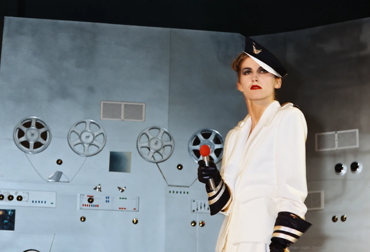 Photographie couleur, format horizontal. Devant une cloison grise constellée de bobines de cinéma, de cadrans et de manettes, un mannequin en uniforme d'officier blanc, casquette marine et gants noirs, tient un micro rouge comme un sceptre. Le décor de science-fiction transforme le podium en salle de contrôle : au Mirano, un défilé n'était jamais qu'un prétexte à raconter une histoire.