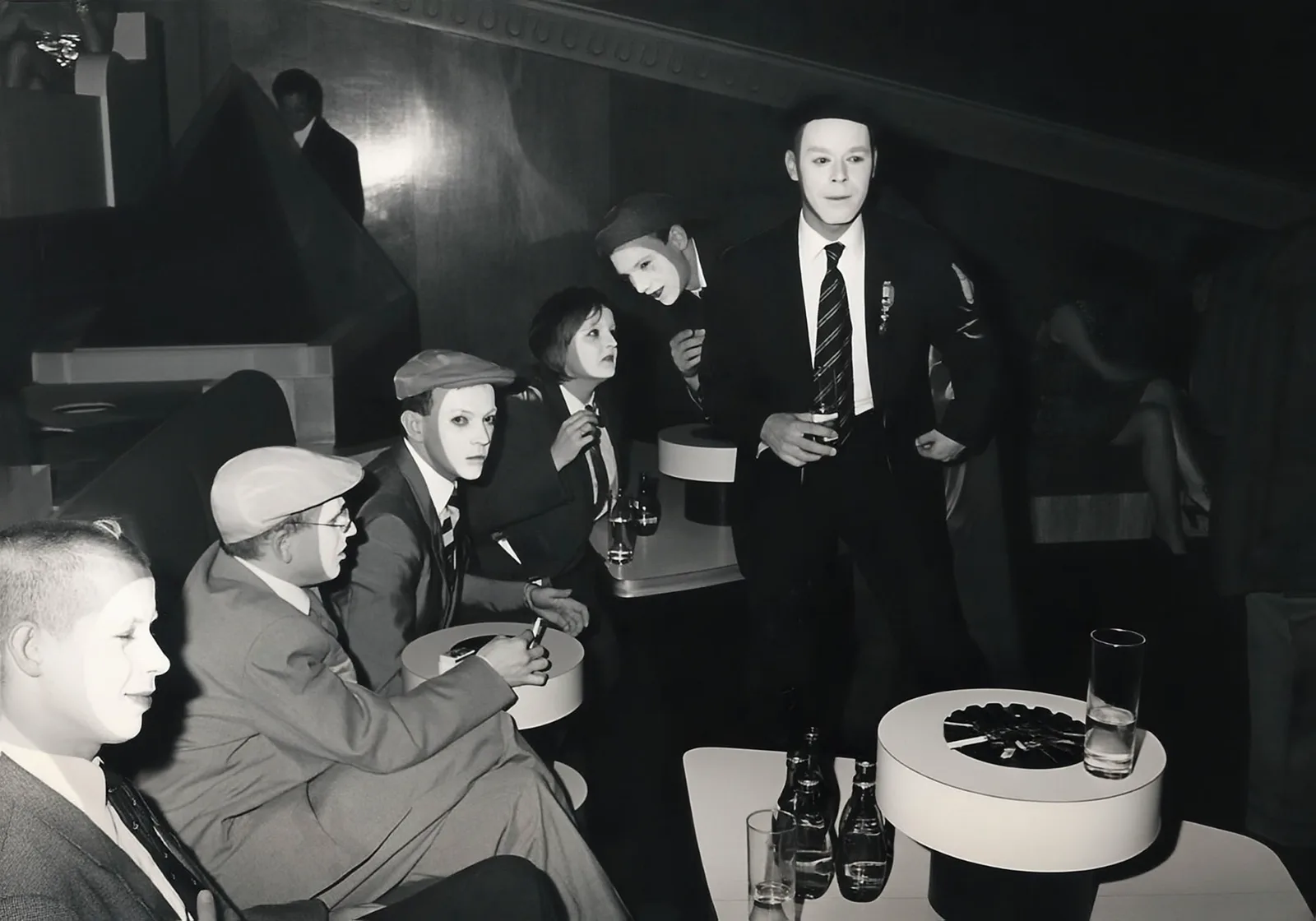 Photographie noir et blanc, format horizontal. Autour des tables rondes du Mirano Continental, l'équipe au complet s'est grimée en personnages d'un film muet des années folles : casquettes, costumes trois pièces, cravates rayées, visages blanchis au fard. Debout, Bruno Bulté, verre à la main.