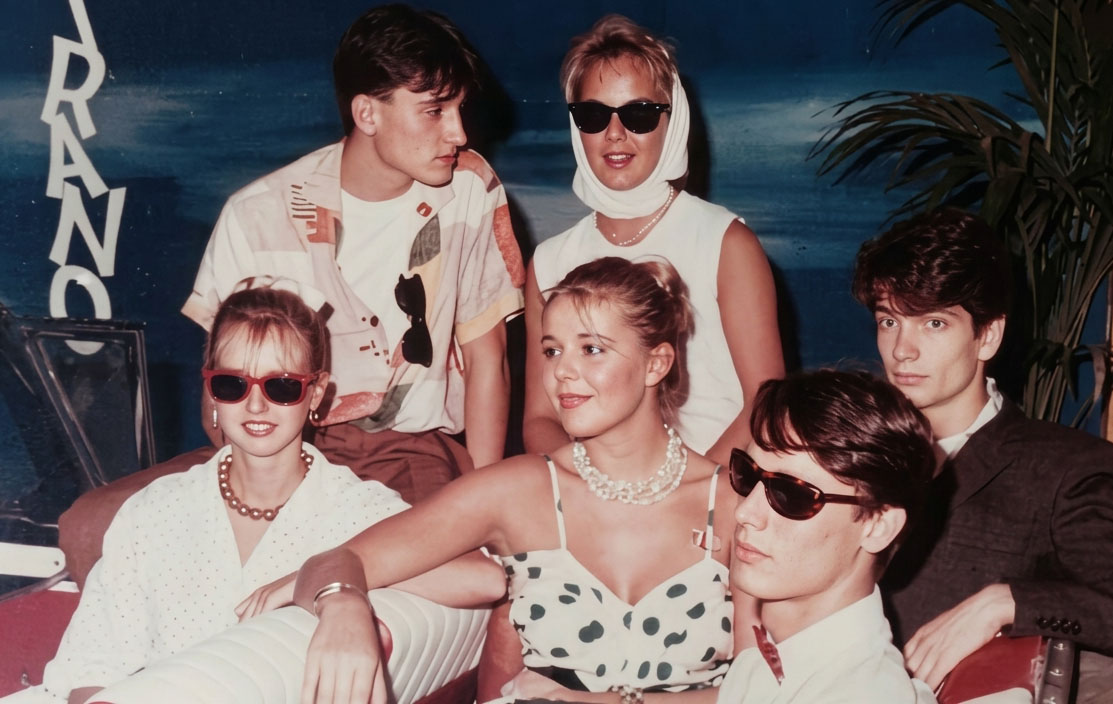 Photographie couleur, format horizontal. Groupe d'invités posant autour de la Cadillac blanche dans le hall d'entrée du Mirano Continental, lunettes de soleil et tenues estivales, devant le décor bleu nuit et le logo vertical du club. 1989.