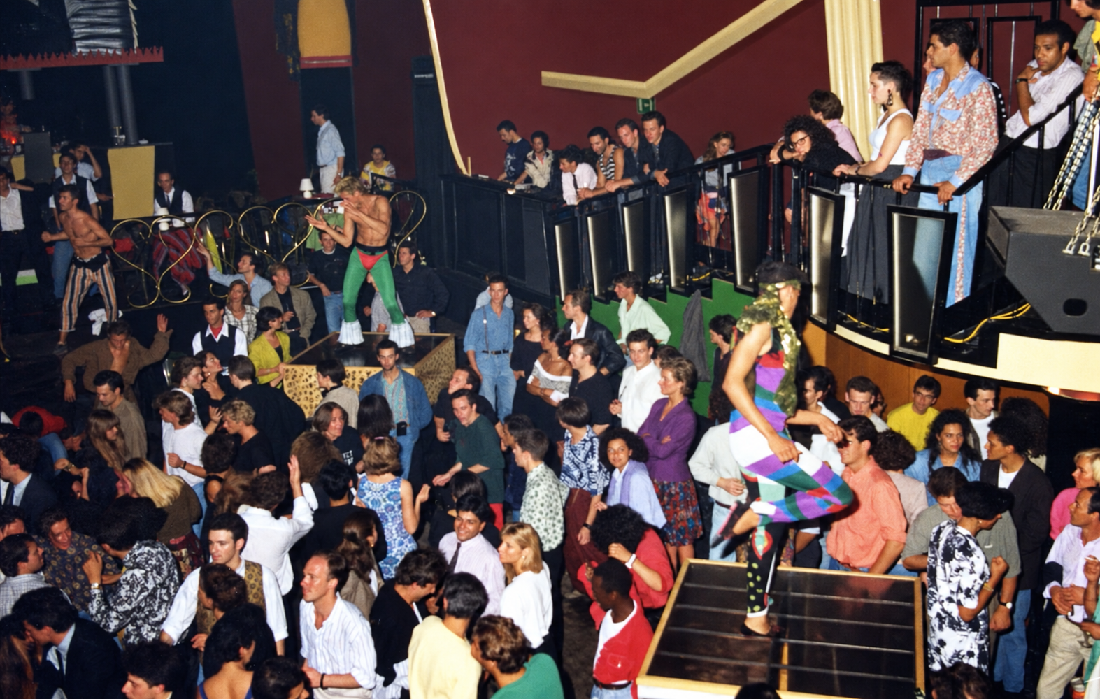 Photographie couleur, format horizontal. Des artistes en costume évoluent au-dessus de la foule du Mirano Continental, entre balcons et escaliers, dans l'effervescence d'une soirée à thème des années 1980.