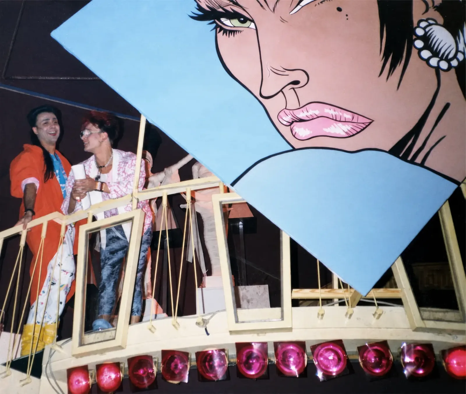Photographie couleur, format horizontal. Au Mirano Continental, en 1986, deux invités se tiennent sur une plate-forme à balustrade blanche, au-dessus d'une rangée de projecteurs roses. Derrière eux, un immense panneau bleu en losange porte un visage de femme géant dessiné comme une planche de bande dessinée : décor de la soirée de lancement de l'album « Bédé Musk » de Jean-Louis Boccar et Patryck de Froidmont, et de leur disque « Looking for the Musk ».