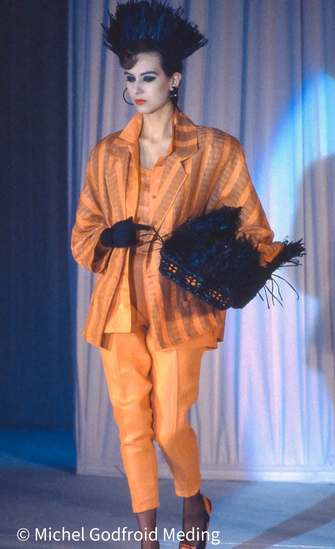 Photographie couleur, format vertical. Une mannequin en ensemble orange safran, veste rayée et pantalon fuselé, chapeau haut en plumes noires et manchon assorti, gants noirs, avance sur le podium devant un rideau de lumière froide. Le contraste des teintes chaudes et du fond bleuté donne à la silhouette une présence d'affiche.