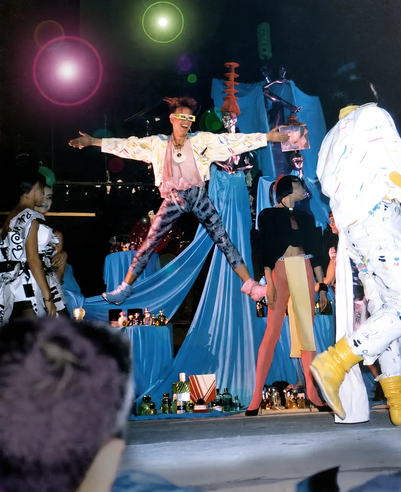 Photographie couleur, format vertical. Sur la scène du Mirano Continental en 1986, un performeur en lunettes jaunes, veste blanche imprimée de graffitis, chemise rose et legging tie and dye saute bras écartés, en plein éclat de rire. Autour de lui, d'autres interprètes en tenues peintes à la main, collants roses et bottes jaunes traversent le plateau, devant un décor de voilages bleus, de mannequins argentés et d'une table couverte de flacons. Deux halos, rose et vert, marquent la photographie.