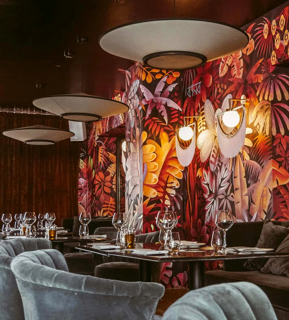 Alignement de tables sous trois larges abat-jour blancs. Sur toute la hauteur du mur, la jungle peinte mêle rouges, roses, ocres et blancs ; deux appliques à franges viennent l'éclairer par l'intérieur.