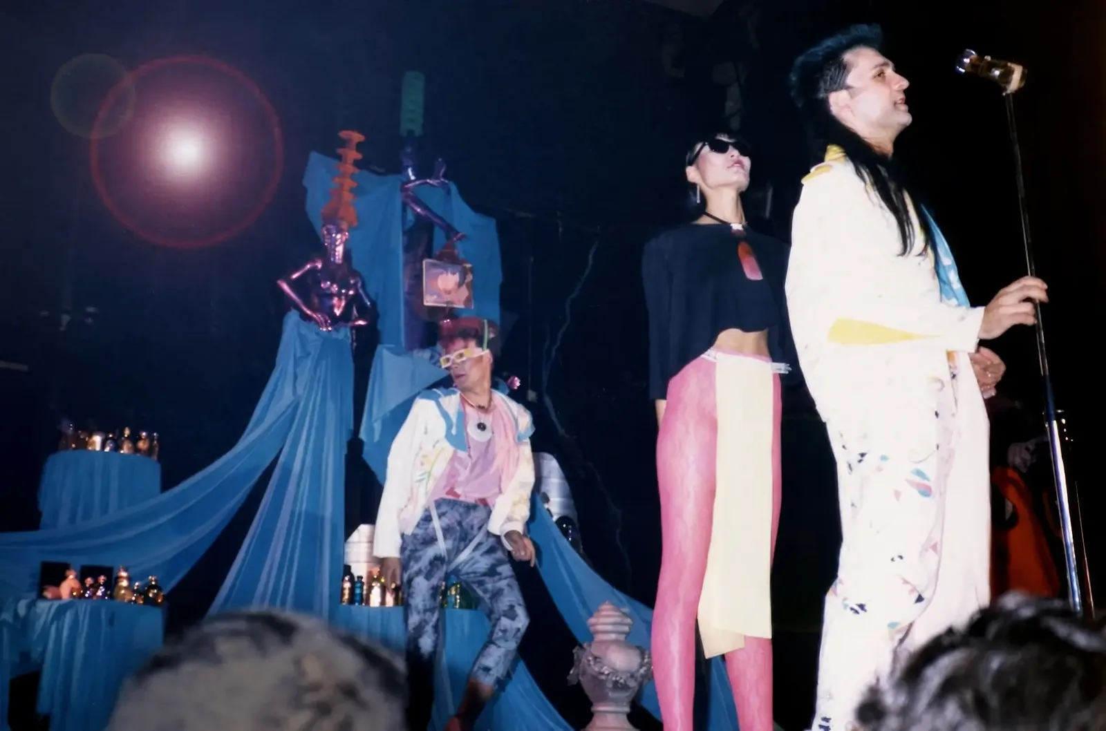 Photographie couleur, format horizontal. Sur la scène du Mirano Continental en 1986, un chanteur en kimono blanc peint tient le pied de micro, une comédienne en lunettes noires et collants roses se tient à ses côtés, un troisième interprète en veste blanche et pantalon marbré danse devant des voilages bleus et une statue laquée. Sur scène, le groupe Les Indians interprète « Looking for the Musk », la bande originale de l'album BD de Jean-Louis Boccar et Patryck de Froidmont.