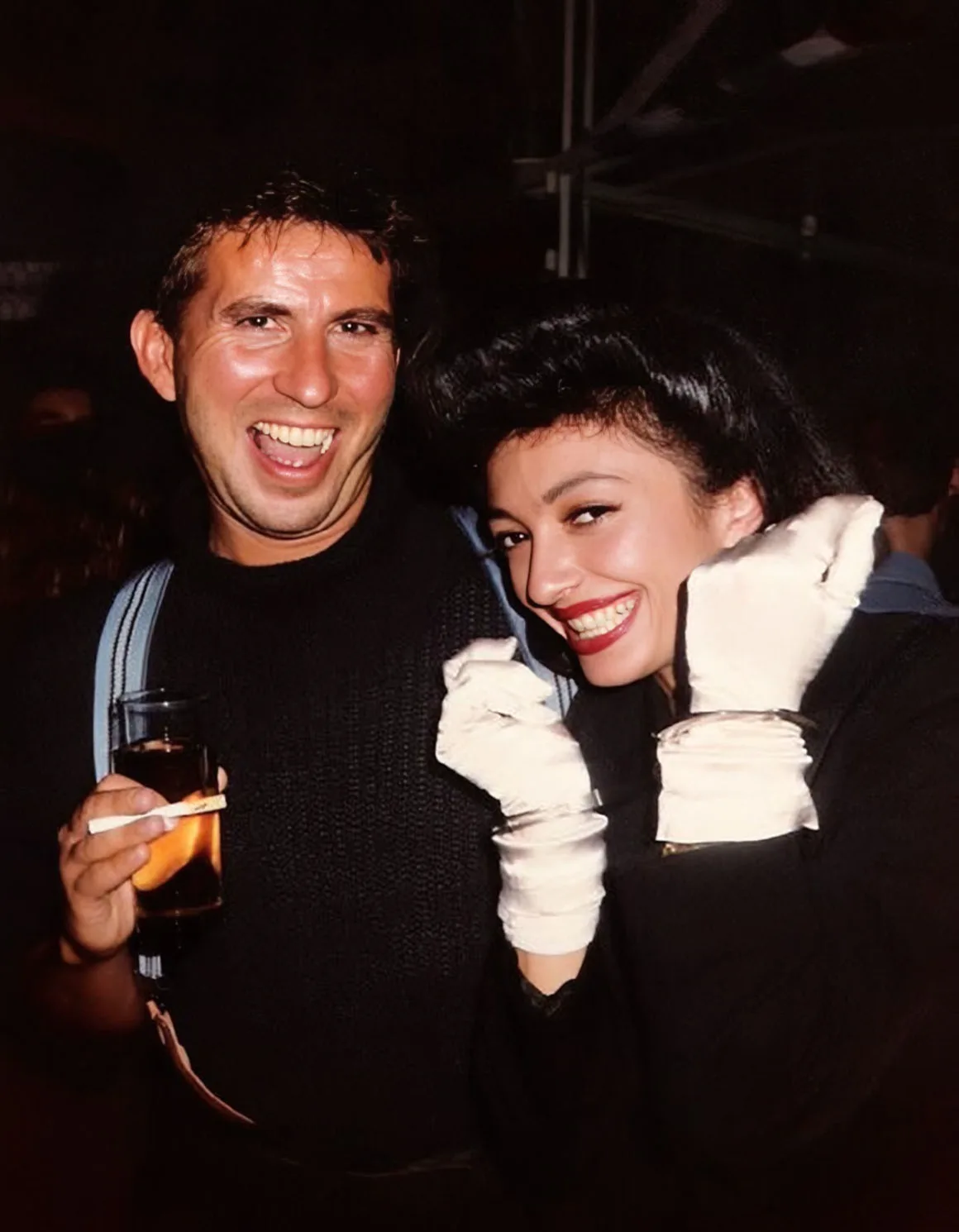 Photographie couleur, format vertical. Caridan, coiffeur attitré des nuits miranistes, verre à la main, pose aux côtés de Filippa Russo, gants blancs et robe noire. Sourires complices au cœur de la nuit. Années 1980.