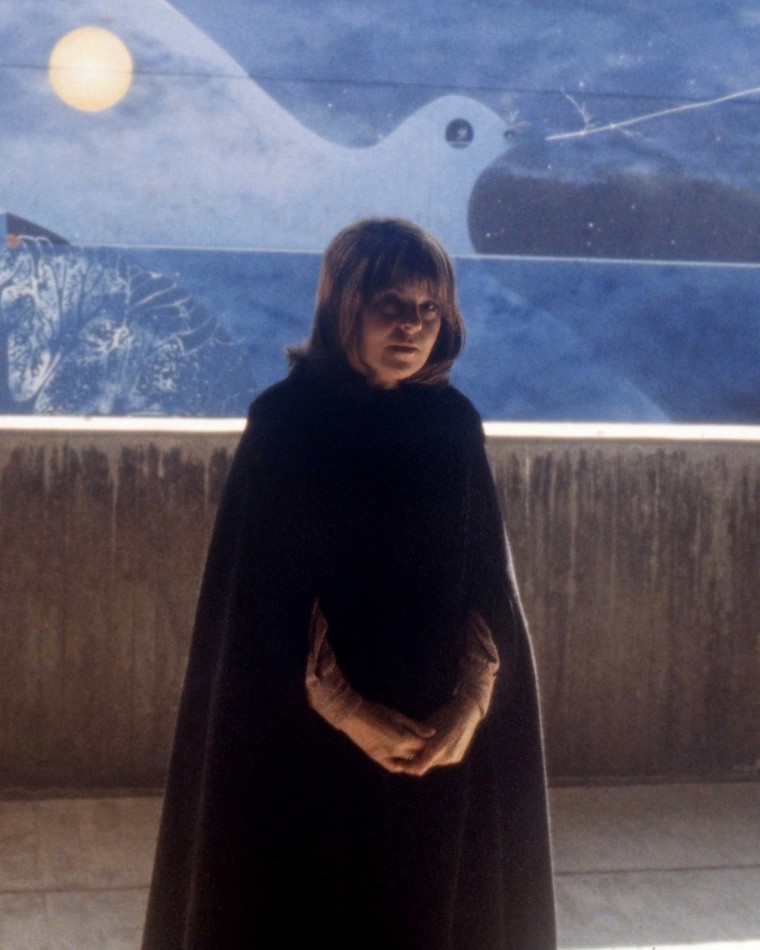 Charline Mahy, « Itinéraire secret d'un point de couleur à travers l'espace », 1978, peinture murale, environ 102 m², Louvain-en-Woluwe (UCL), Bruxelles. L'artiste pose devant l'œuvre : le bandeau peint, bleu nuit ponctué d'un disque solaire et d'une grande forme claire, contraste avec le béton brut du muret au premier plan.